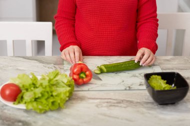 Pişirme zaman kavramı. Kırpılmış görüntü hamile olmak taze sebze salatası hazırlar, salatalık, biber ve domates bıçakla keser, mutfak masasına teşkil etmektedir, kırmızı sıcak kazak giyer. Beslenme kavramı