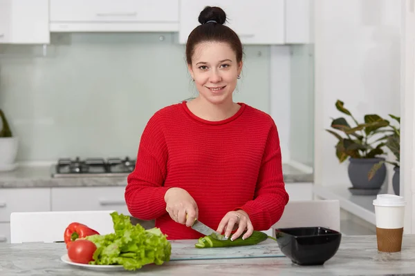 肯定的なブルネット妊婦夕食にサラダを作る キュウリをナイフでカット カジュアルな服を着ている 健康的な栄養には 台所のテーブルのそばに立ちます 健康食品 料理の概念 — ストック写真