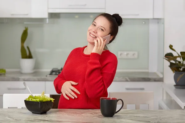 幸せなブルネット妊娠中の母親が携帯電話を介して良い会話 おなかの上の手を保持 ベジタリアン サラダを食べる ホットド リンクを飲む友人とニュースを共有 キッチン インテリアに対するポーズ — ストック写真