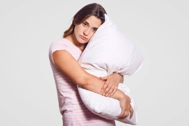 Yatay atış ciddi üzgün kadın beyaz yastık yakından taşır, pijama giyiyor, doğrudan kameraya benziyor, beyaz arka planı teşkil etmektedir, dinlenmeye, uyku için hazır yemek istiyor, tatlı rüyalar vardır