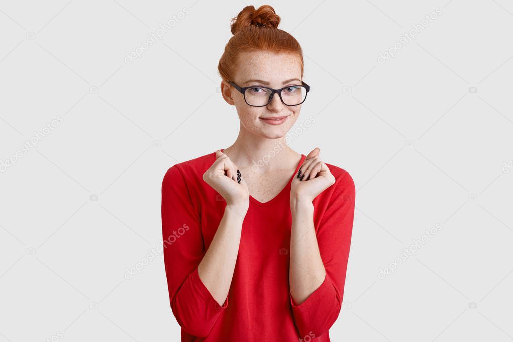 Happy red haired freckled young woman with knot keeps hands pressed, waits for something with intrigue, looks through transparent glasses, dressed in red clothes, isolated over white background