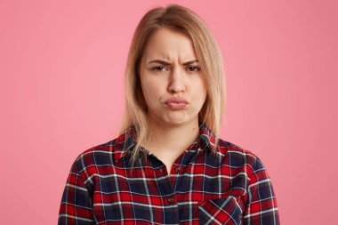Memnun sarışın kadının kapalı atış dudaklar Gücenmek ve yüz frowns, gibi bir şey değil, somurtkan yüz ifadesi olan, pembe arka plan üzerinde izole kareli gömlek giyer, memnun yüz buruşturma yapar