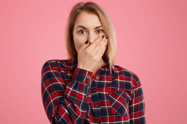 Korkmuş güzel kadın ağız kapsar, korku dolu gözlerle bakar, kısa sarı saçlı, pembe bir arka plan üzerinde izole damalı şık gömlek giyer, surprisement ve güvensizlik ifade