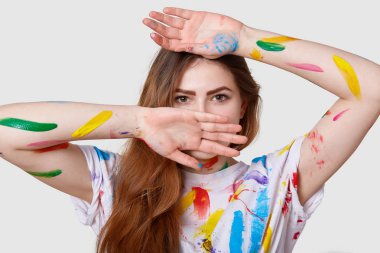 Headshot olan çekici ciddi sevimli kadın koyu renk gözleri vardır, beyaz arka plan üzerinde izole renkli akrilik boya ile lekeli elleriyle yüzünü kapsar. İnsanlar, yaratıcılık ve işgal kavramı