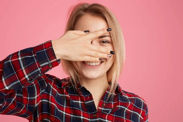 Photo of pleasant looking blonde European female peeks through fingers, has toothy smile, expresses happiness, hides face from someone, models against pink background. Horizontal studio shot