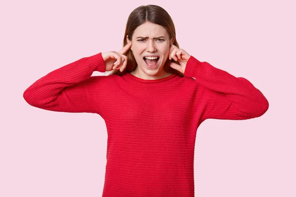 照片中 绝望的高加索妇女用手指插断耳朵 愤怒地喊道 穿着红色的套头衫 被烦人的声音激怒 被浅粉色背景隔离 表达负面的感情 — 图库照片