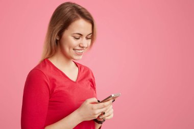 Gülümseyen hoş görünümlü kadın kırmızı kıyafeti akıllı telefon, arama sosyal ağlar, kısa saç modeli, kablosuz internet, metin için boş alan ile pembe arka plan üzerinde izole bağlı olan tutar