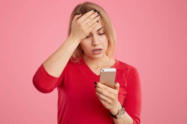 Stresli genç bayan alnına palm tutar, internet, cep telefonu odaklı şok haber okur, pembe arka plan üzerinde izole kırmızı rahat jumper giyer. Baş belası kadın pozlar kapalı.