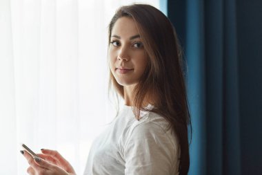 Çekici Bayan siyah saçlı görünüyor doğrudan ile kapalı atış beyaz t gömlek giymiş kamera cep telefonu tutan, metin mesajı gönderir, pencere perdeleri, kablosuz Internet bağlantısı ile yakın duruyor