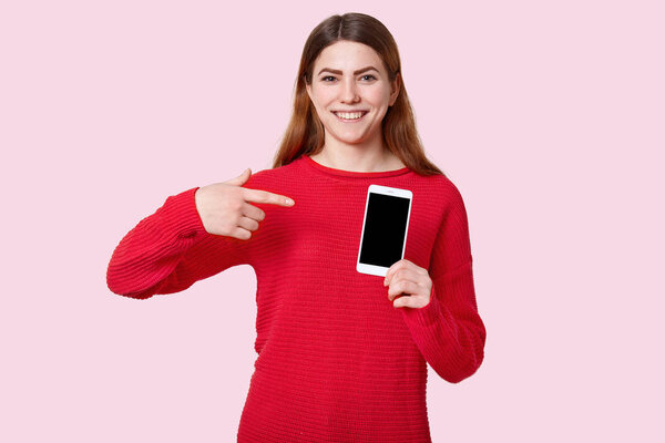 Посмотри на этот гаджет. Счастливая улыбающаяся европейская молодая женщина указывает на современный мобильный телефон с пустым экраном для вашего рекламного контента, одетый в негабаритный красный свитер, позирует на розовом фоне
