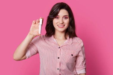 Dalgalı saçlı, gülümsüyor şirin pozitif kadın geniş, dudak parlatıcısı, yeni kozmetik üründen memnun tutar, pembe şık gömlek giyer, pembe bir arka plan karşı modelleri. Feminity ve güzellik kavramı