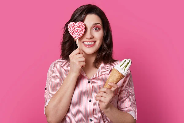 Positive young cute lady covers eye with sweet candy, holds ice cream in other hand, has fun indoor, being sweet tooth, dressed in striped shirt, isolated over pink background, has glamour look