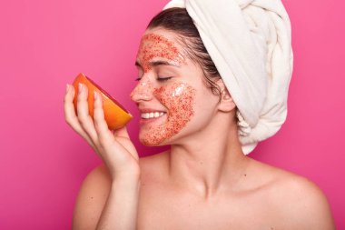 Neşeli genç kadın modeli kozmetik temizlik maskesi yüzünde, vardır kokuyor greyfurt, yan duruyor, havlu kafasına sarılmış, çıplak omuz, sağlıklı cilt, pembe bir arka plan üzerinde izole gösterir