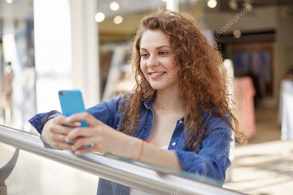 Young curly beauty stands in shopping mall, hold telephone, phone in pleasant color case. Person expresses joy because, comes message where she saw salary of her work in fashionable model agency.