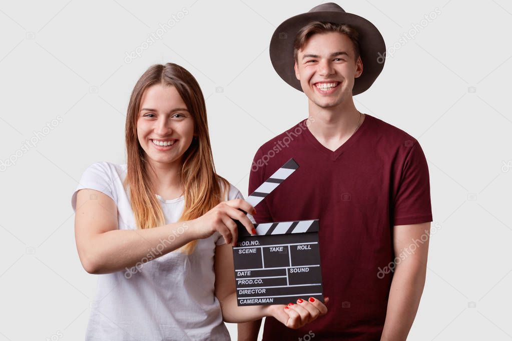 Happy joyful couple stand closely, hold clapperboard in front, prepare for next scene and take, expresses positive emotions in cinema, isolated over white background. Slate film, movie production