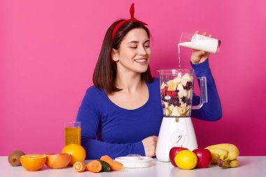 Güzel gülümseyen kız smoothies sabah yemek yapıyor. Birçok meyve masada. Sevimli kadın süt blender için ekler. Güzel erkek coktail yapar. Sevimli Bayan pozlar Studio gül duvara.