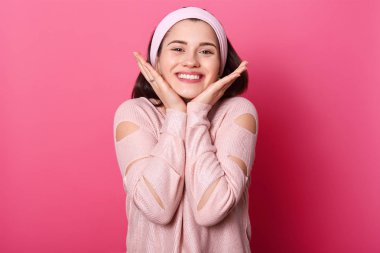 Mutlu güzel kız gülmek görünümü kamera ile yanaklarda ellerini tutar. Genç Bayan iyi hissettiriyor. Sevimli kadın seviyor adam görür. Modeli pozlar pembe duvar moda gömlek ve hairband karşı.