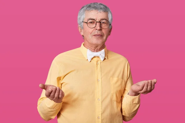 Varón anciano no sabe qué tipo de emoción expresar, levanta las palmas. Viejo con camisa amarilla y gafas redondeadas negras siente cierta confusión. Senior no entiende lo que pasó . —  Fotos de Stock