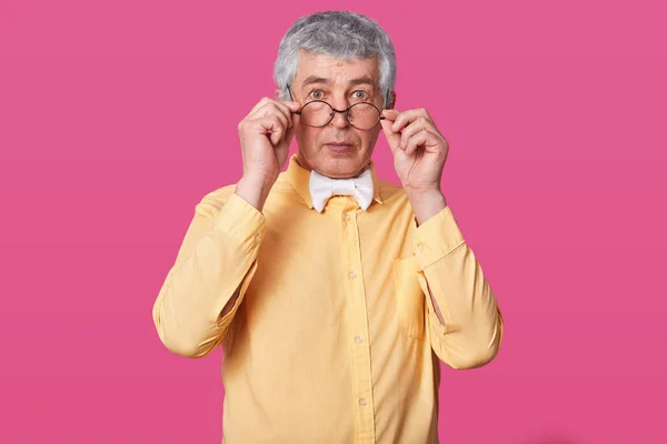 Man with black rounded glasses on tip of nose. Suprised old male holds spectacles with hands. Eldery senior with wide opened eyes hears shocked information isolated over pink background. — Stock Photo, Image