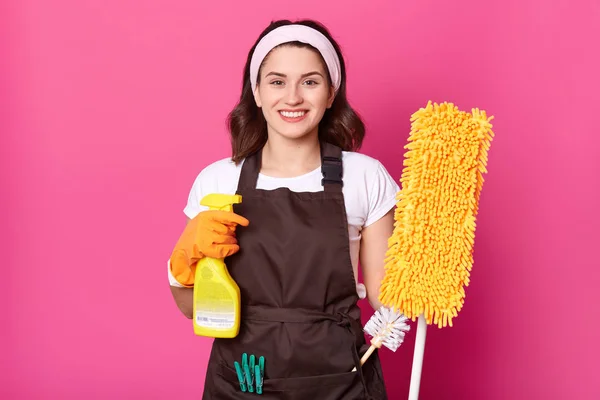 Cheerful young housewife wears white t shirt, brown apron with green clothespins, orange rubber gloves for hands protection. Holds spray and mop. Smiling female cleans house. Works at home. — Stock Photo, Image