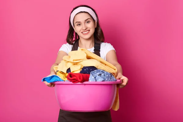 Young woman holds basin full of clean linen. Beautiful housewife looks happy after doing laundry. Smiling female works about house. Maid in brown apron enjoys fresh colourful linen on pink background. — Stock Photo, Image