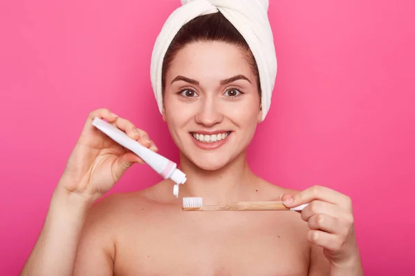 Mujer joven desnuda con toalla blanca en la cabeza lista para cepillarse los dientes. Mujer sonriente pone pasta de dientes en el cepillo de dientes. Linda chica con sonrisa perfecta se ve feliz en el baño. concepto de estilo de vida saludable . — Foto de Stock