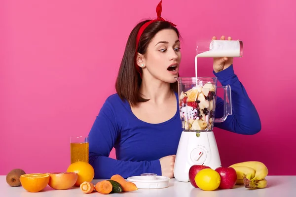 Young woman making healthy smoothie. Slender female with opened mouth adds milk to blender. Suprised girl dresses shirt and red hairband against rose wall. Model poses in studio. Lifestyle concept.