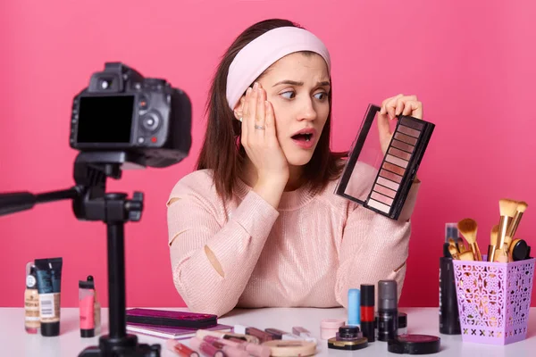 Encantadora joven se sienta frente a la cámara, ha sorprendido a la expresión, rodeado de productos de belleza. La chica sostiene la sombra de ojos, mantiene su mano en la mejilla. Vlogger aislado sobre fondo rosa . — Foto de Stock