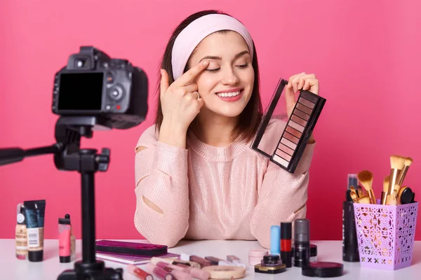 Atractiva hembra se sienta delante de la cámara fija en el trípode. Chica bonita mira la sombra de ojos que sostiene en la mano. El exitoso bloguero hace video. Señora tiene expresión alegre aislado sobre fondo rosa . — Foto de Stock