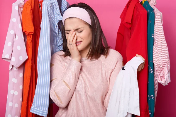 Crying woman keeps right hand near her face, holds hanger with white shirt while standing at her wardrobe. Dark haired woman has nothing to wear and no money for buying new outfit. People, problems.