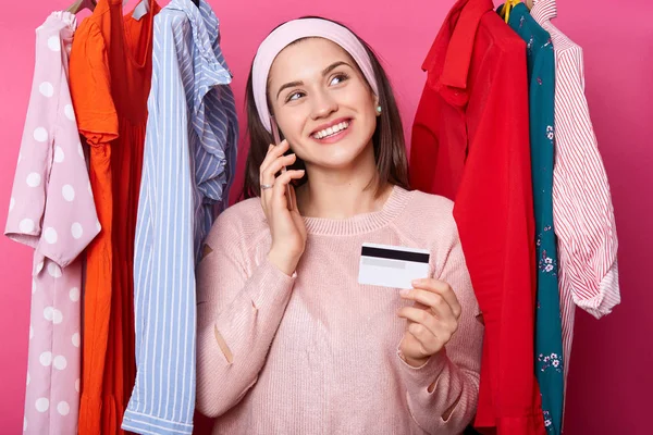 Junge Frau mit Kreditkarte träumt vom Einkaufen, während sie in der Nähe vieler Kleiderbügel mit bunten Kleidern vor rosa Hintergrund steht. fashoin male mag Online-Shopping. lächelndes Mädchen kauft Kleid. — Stockfoto