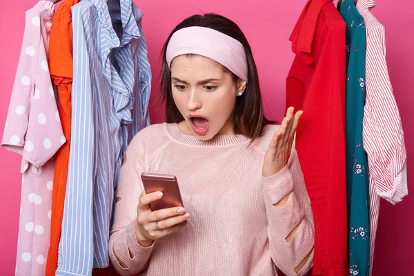 Shocked woman looks at smartphone display. Interesting information on screen. Dark haired lady raises palm and looks surprisingly at her gadget. Lots clothes behind beautiful female in shopping mall. — Stock Photo, Image