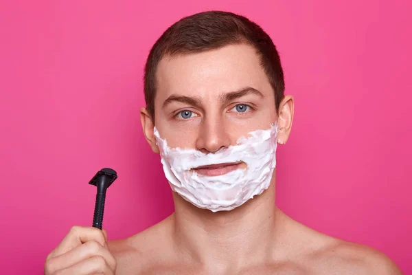 Ung attraktiv kille som är redo för rakning med rakhyvel i badrum, sätter kräm på ansiktet, över rosa bakgrund. Stilig man med skum på hans kinder poses i studio. Härdat hane med naken överkropp. — Stockfoto