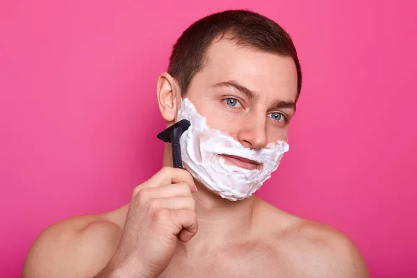 Close up of handsome shirtless man stands isolated over pink background. Guy with naked shoulders holds razor. Boy shaves and does everyday morning procedures. Skin care and lifestyle concept. — Stock Photo, Image