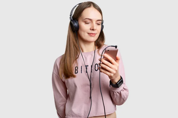 Adegan dalam ruangan gadis remaja yang difokuskan pada layar telepon pintar, mendengarkan musik dengan headphone dari playlist, menikmati suara murni keras, memiliki rambut panjang, terisolasi di atas latar belakang putih. Konsep waktu luang — Stok Foto