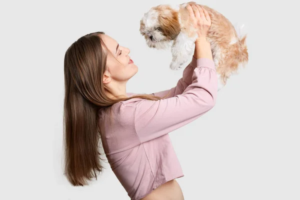 Sideways disparo de mujer de pelo oscuro complacido sostiene a su amigo de cuatro patas en el aire, juega con el perro pedigrí, disfruta del tiempo libre, vestido con la parte superior de moda, aislado sobre fondo blanco. Animales. —  Fotos de Stock