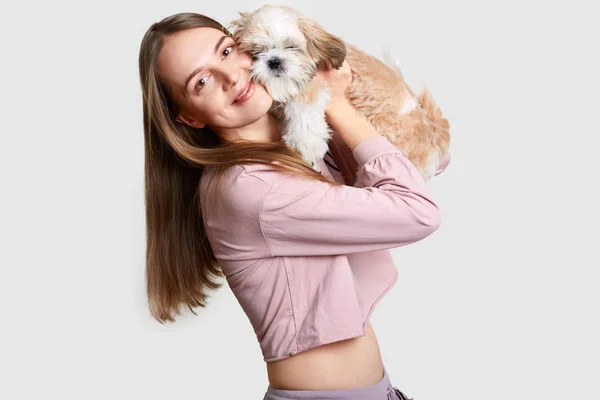 Mujer europea positiva con el pelo largo abraza a su mascota favorita con piel esponjosa, vestida con top casual, muestra el vientre desnudo, estar en buena forma, le gustan los animales, aislado sobre fondo blanco —  Fotos de Stock