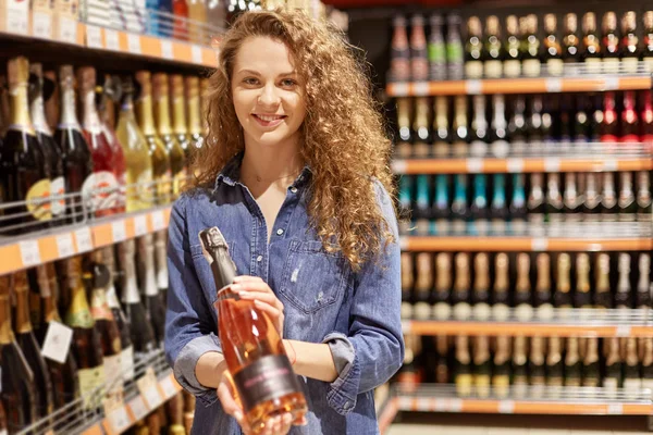 Charming young woman with brown curly hair , in denim clothes. Holds bottle of alcoholic drink, stands near supermarket shelves , finds wine or champagne . Smiling girl buys necessary drink for party. — Stock Photo, Image