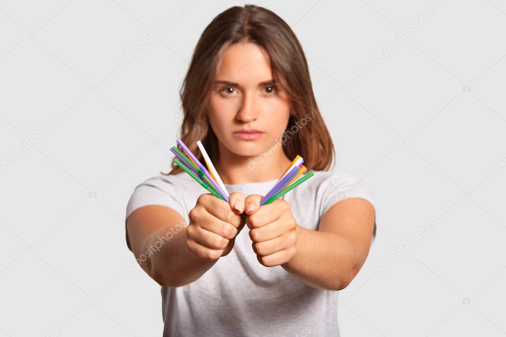 No plastic pollution concept. Beautiful serious self confident female presses plastic straws in hands, being against of pollution of environment, dressed in casual t shirt, isolated over white wall