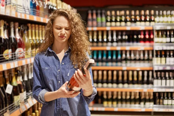 Attractive young female with curly hair, wears demin jacket. Holds bottle of alcoholic drink, reads information about wine, looks unhappy. Beautiful girl finds champagne. Woman chooses wine in mall. — Stock Photo, Image