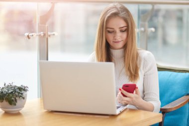Konsantre kadının fotoğrafını modern teknolojiler kullanır, açılan dizüstü bilgisayar önünde oturur, cep telefonu, surfes sosyal ağlar, arkadaş, pozlar Cafe Divan metin mesajı gönderir tutan