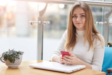 Memnun güzel bayanı rahat kıyafetler içinde ileti göndermek için cep telefonu kullanır, kahve masada oturur kapalı dizüstü, güzel yeşil çiçek vazo içinde çağrı erkek için bekler.