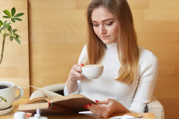 Concentrated pretty young woman reads book with cup of cappuccino, focused on reading, enjoys interesting exciting novel, poses in cafeteria, dressed in white sweater, has spare time after studying. — Stock Photo, Image