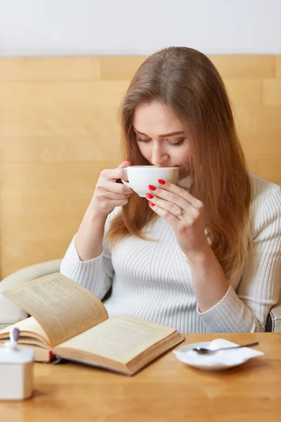 Tiro vertical de mulher encantadora despreocupada bebe chá quente de caneca branca, lê história fantástica, gosta de livro, posa no café, espera por alguém, tem hobby, gosta de bebida aromática. Conceito de lazer — Fotografia de Stock