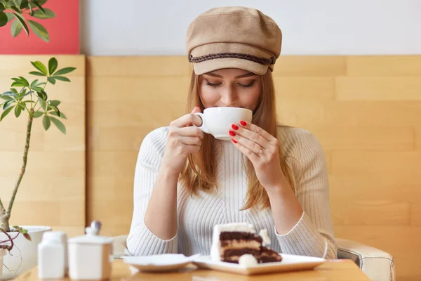 Jovem estilista bonita elegante tem pausa após o trabalho, visita cafetaria local, bebe bebida quente, segura caneca branca, come bolo muito saboroso, usa chapelaria elegante e jumper casual — Fotografia de Stock