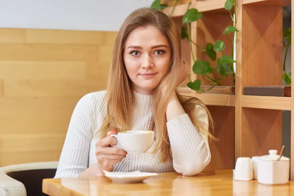 Foto de bela jovem fêmea de aparência europeia, vestida com roupas brancas, bebe café quente aromático ou chá na cafetaria acolhedora, olha diretamente para a câmera, tem bom descanso durante o fim de semana . — Fotografia de Stock
