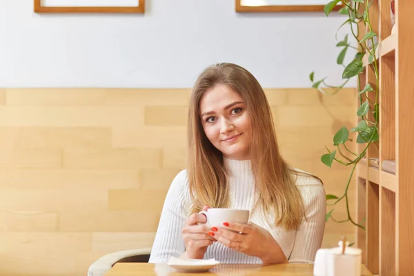 Imagem de modelo feminino jovem encantador com cabelo claro reto, detém caneca de bebida, tem tempo livre, encontra-se com o parceiro no café, goza de atmosfera calma, gosta de beber café. Hora de almoçar — Fotografia de Stock