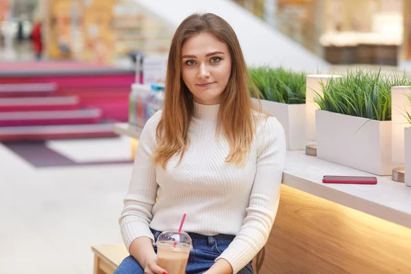 Piacevole cercando giovane donna vestita con maglione e jeans, beve milkshake, si siede nel centro commerciale contro il verde, smart phone vicino, aspetta qualcuno, ha fine settimana, va a fare shopping. Ragazza con bevanda — Foto Stock