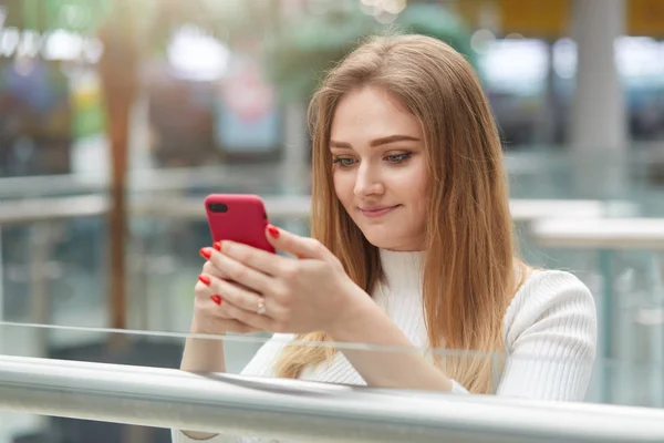 Pretty lovely young woman with makeup, has long hair, dressed in casual jumper, holds smart phone, makes shopping online, checks bank account, stands in shopping mall against blurred background — Stock Photo, Image