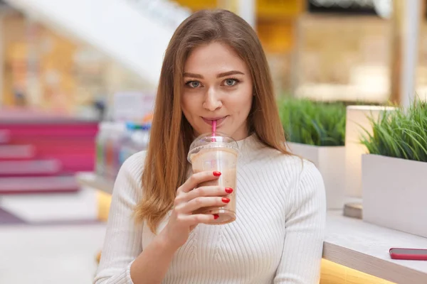 Menina linda olha para a câmera com expressão satisfeita, bebe coquetel de leite, usa saltador branco, tem maquiagem, posa contra o interior borrado café, goza de tempo livre. Peope, conceito de bebida — Fotografia de Stock